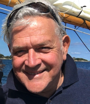 HEADSHOT PHOTO OF WRITER ROBERT F. GODWIN ON A SAILBOAT, SUNGLASSES UP ON HIS FOREHEAD, THE BOOM AND SAIL OF THE SAILBOAT IN THE BACKGROUND AGAINST A BLUE SKY WITH THE COAST OF CHILE IN THE BACKGROUND